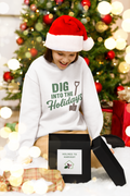 Person wearing a 'DIG INTO THE HOLIDAYS' sweater with a black gift box in front of a decorated Christmas tree.