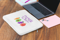 White pouch with colorful designs and text on a wooden surface with a laptop and notebook.