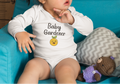 Baby wearing a 'Baby Gardener' onesie sitting on a blue couch with a teddy bear.
