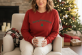 Woman wearing a red 'Merry Christmas' sweater holding a mug in a cozy living room with a Christmas tree.
