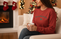 Woman wearing a red 'Merry Christmas' sweater sitting on a couch with a fireplace and decorated tree in the background.
