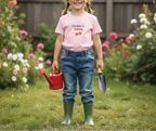 Child in a garden holding a red watering can and a shovel, wearing a pink t-shirt with text.