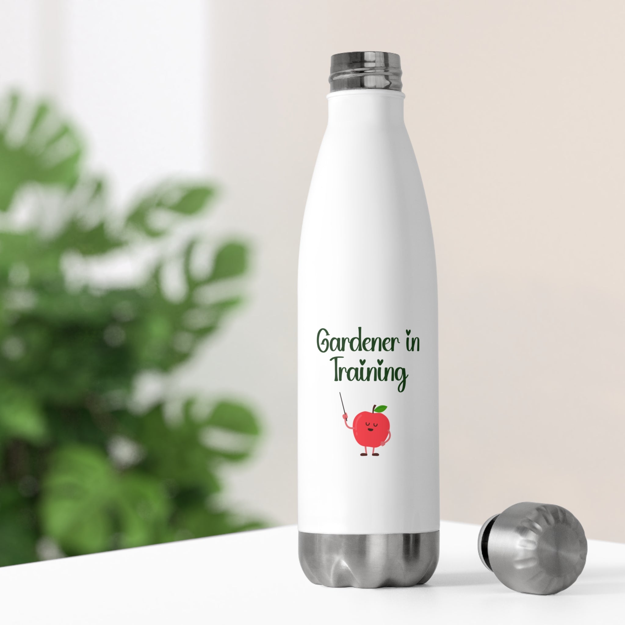 White water bottle with 'Gardener in Training' text and apple graphic on a blurred green plant background