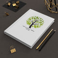White notebook with green tree design and text on a dark surface with office supplies.