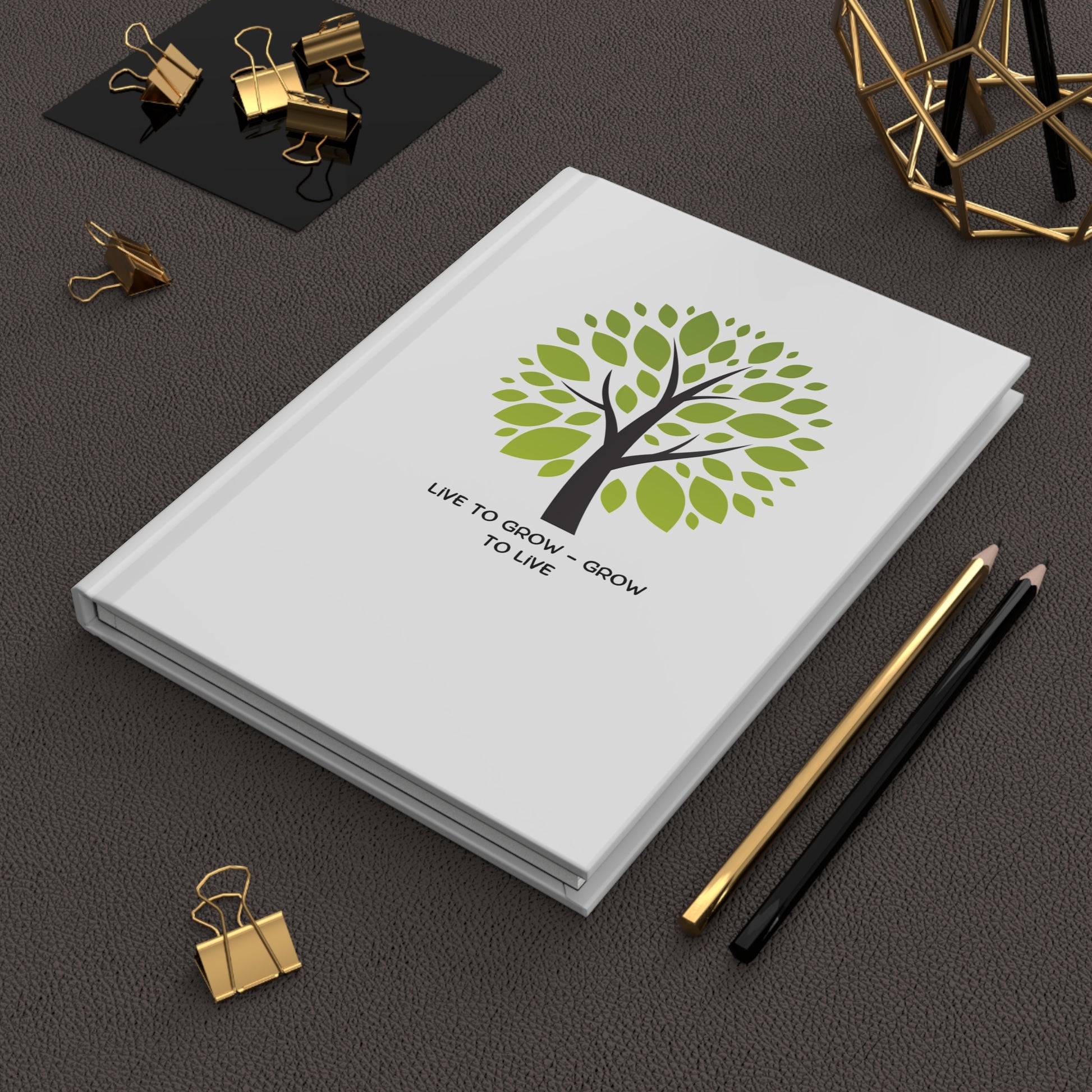 White notebook with green tree design and text on a dark surface with office supplies.