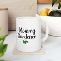 White mug with 'Mommy Gardener' text on a coaster, surrounded by plants and a wooden surface.