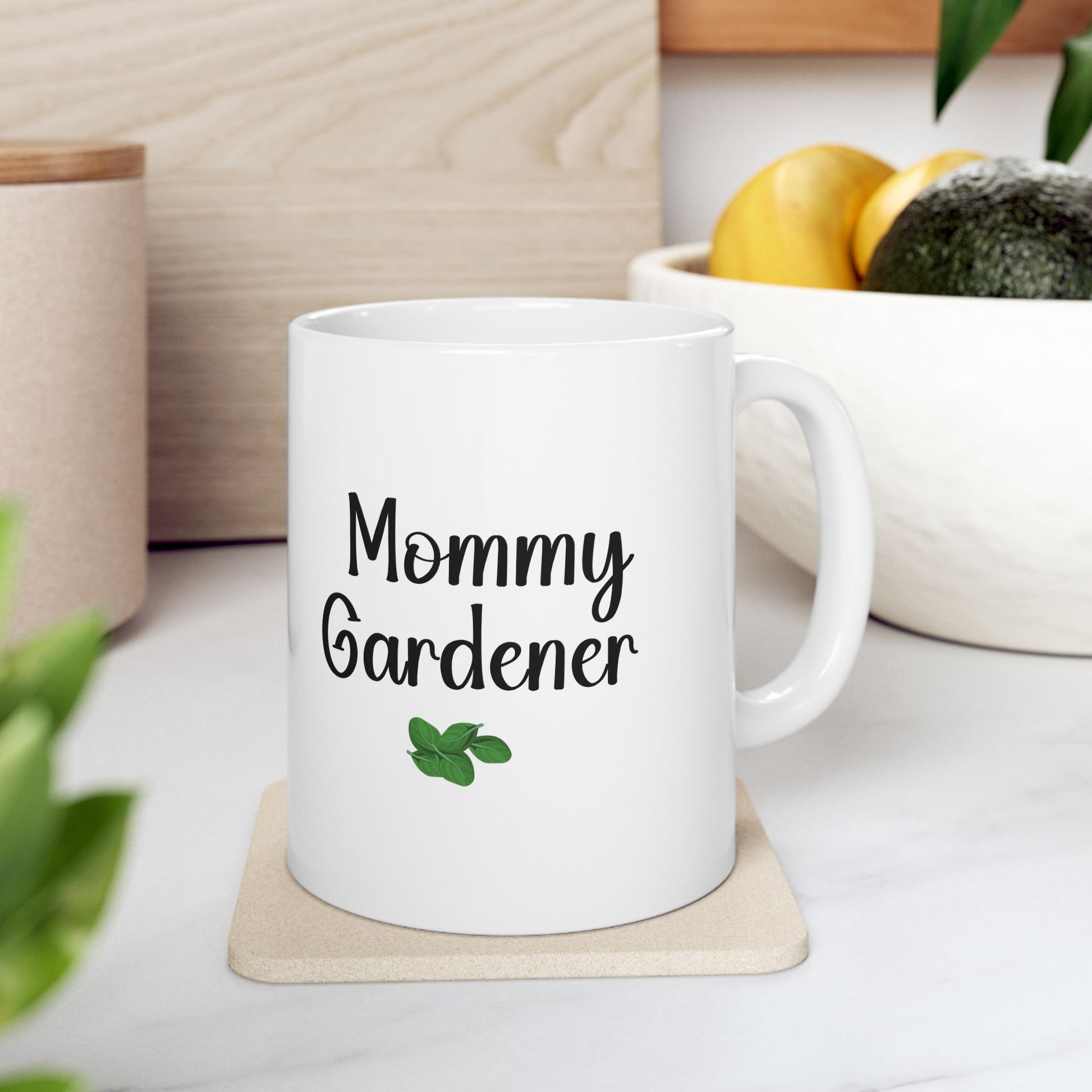 White mug with 'Mommy Gardener' text on a coaster, surrounded by plants and a wooden surface.