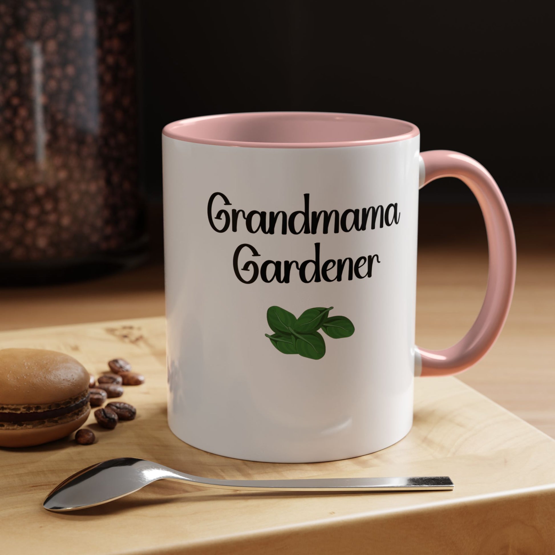 White mug with pink interior and handle, featuring 'Grandma Gardener' text and a leaf graphic, on a wooden surface with coffee beans and a spoon.