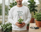 Man wearing a white sweatshirt with 'Gardening is My Therapy!' holding a potted plant in a greenhouse.