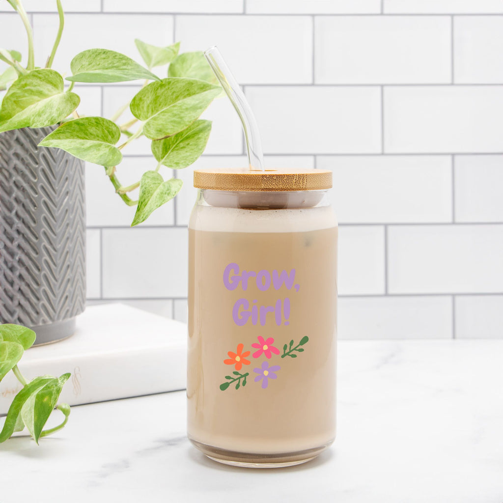 Candle in a glass jar with a wooden lid and colorful text and graphics on a white surface with a plant in the background.