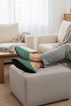 Person wearing green slippers on a beige ottoman in a living room.
