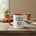 White mug with red interior and handle, featuring 'Gardening Grandpa' text and heart design on a wooden table with gardening tools and plants.