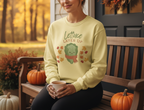 Person wearing a light yellow sweatshirt with a lettuce graphic and text, sitting on a wooden bench with pumpkins and autumn decorations.