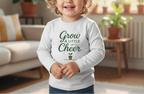 Child wearing a white sweatshirt with 'Grow a Little Cheer' text in a home setting