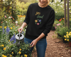 Person holding a watering can in a garden wearing a black sweatshirt with colorful floral design and text.