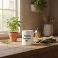 White mug with 'Mommy Gardener' text held by a hand with red nail polish on a white background