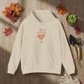 Beige hoodie with 'Fall into Gardening' design on a wooden surface with autumn leaves and gardening tools.
