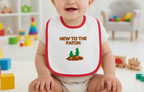 Child wearing a bib with 'New to the Patch' text and cactus graphic in a playroom setting.