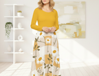 Woman wearing a yellow top and floral skirt in a bright room with shelves and artwork.