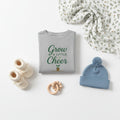Gray long-sleeve shirt with green text 'Grow a Little Cheer' on a beige background
