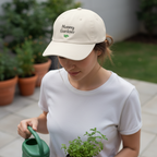Woman wearing a 'Mommy Gardener' cap holding a plant and watering can outdoors.