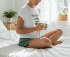 Person sitting on a bed holding a mug with a 'Wake Up & Garden' shirt.
