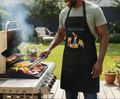 Man grilling outdoors wearing a black apron with colorful graphics and text.