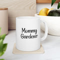 White mug with 'Mommy Gardener' text on a coaster, surrounded by plants and a wooden surface.
