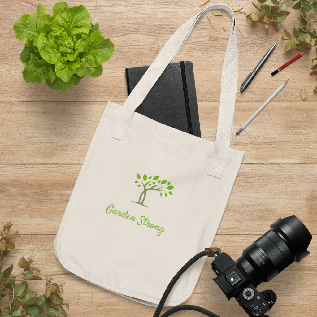 Beige tote bag with 'Garden Strong' logo on a wooden surface with stationery items and plants.