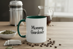 Mug with 'Mommy Gardener' text on a table with coffee beans and tea leaves.