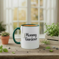 White mug with 'Mommy Gardener' text on a wooden table with plants and a blurred background