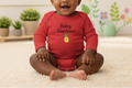 Baby wearing a red 'Baby Gardener' onesie sitting on a carpeted floor with plants and flowers in the background.