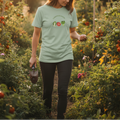 Person walking through a garden wearing a green t-shirt with a logo, holding a trowel.