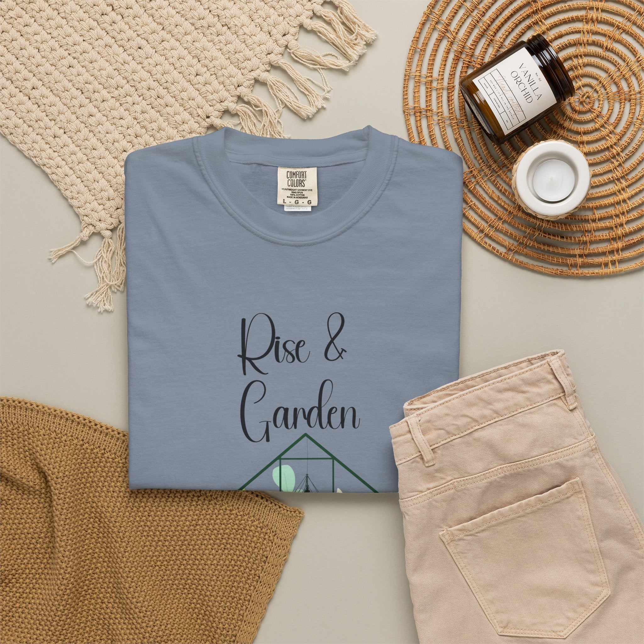 Folded blue t-shirt with 'Rise & Garden' text on a neutral surface with a candle and woven mat.