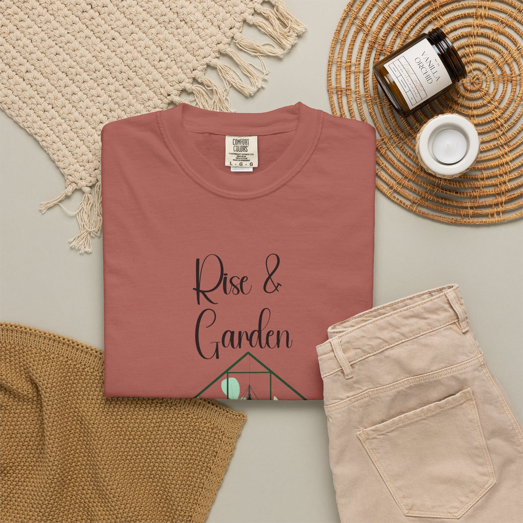 Folded pink t-shirt with 'Rise & Garden' text on a neutral surface with pants and decor items.