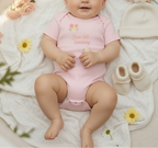 Baby in a pink onesie with floral designs on a soft surface with flowers and shoes.