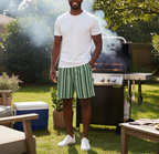 Man standing outdoors by a grill with smoke, wearing a white t-shirt and green striped shorts.