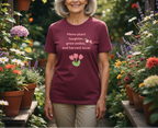 Woman wearing a maroon t-shirt with a floral design and text, standing in a garden.