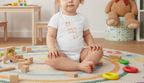 Child sitting on a colorful rug with toys around, wearing a white onesie with text.