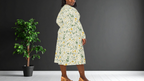 Woman wearing a floral dress standing next to a potted plant against a dark wall.
