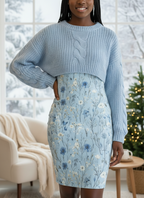 Woman wearing a light blue floral dress in a bright room with plants.