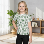 Young girl wearing a white t-shirt with green floral patterns in a room with plants and books.