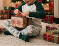Child opening a Christmas present surrounded by wrapped gifts on a carpeted floor.