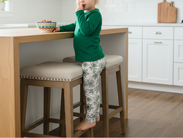Child in a green shirt and patterned pants sitting on a bar stool in a kitchen.