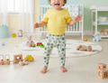 Child in a yellow shirt and floral pants standing in a nursery room with toys and furniture.