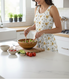 Woman in a kitchen preparing a salad with various ingredients on a white countertop.