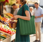 Woman shopping at an outdoor market, holding a basket of grapes and a green striped bag.