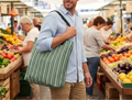 Man holding a striped reusable bag in a bustling market with fresh produce.