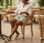Woman sitting at an outdoor cafe table with a drink, wearing a white shirt and floral shorts.