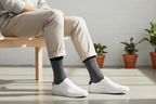 Person wearing white sneakers and dark patterned socks sitting on a wooden stool with plants in the background.